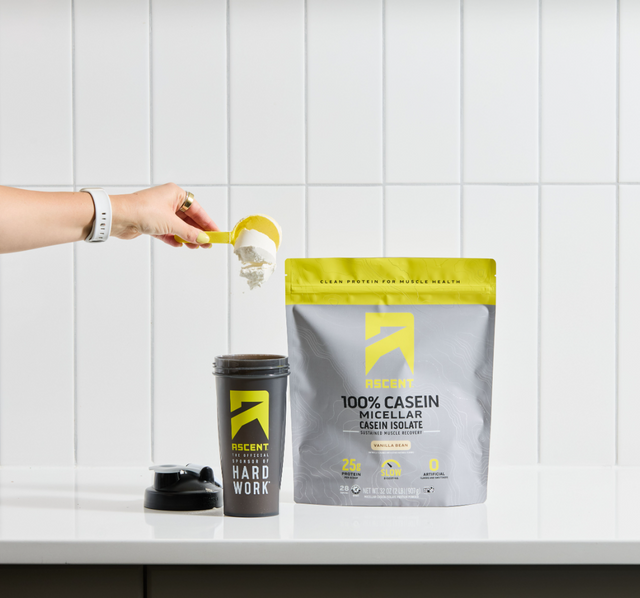 A hand holding a plastic scoop and pouring Micellar Casein powder into a shaker bottle next to a bag of Vanilla Bean Micellar Casein.
